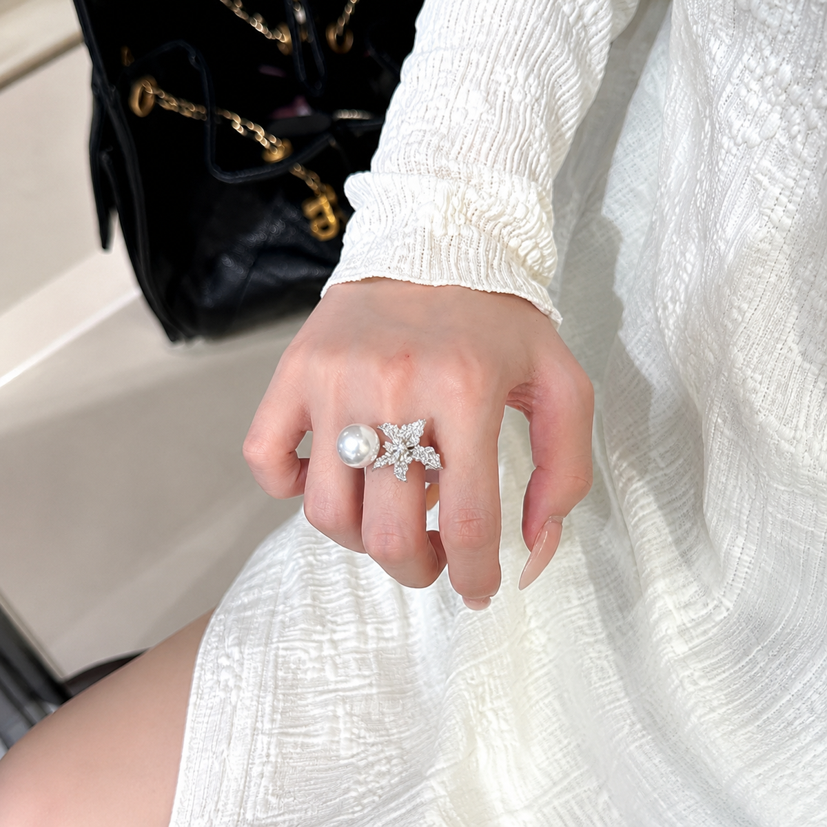A close-up of a hand wearing a silver-toned ring with a large white pearl and a sparkling star-shaped setting, shown against a white textured dress and a black quilted handbag.