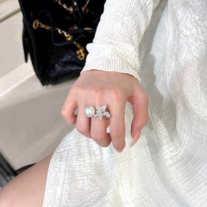 A close-up of a hand wearing a silver-toned ring with a large white pearl and a sparkling star-shaped setting, shown against a white textured dress and a black quilted handbag.