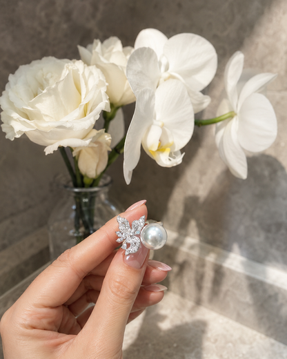 A hand holds a silver butterfly-shaped stud earring with a white pearl, with white flowers in a glass vase blurred in the background.