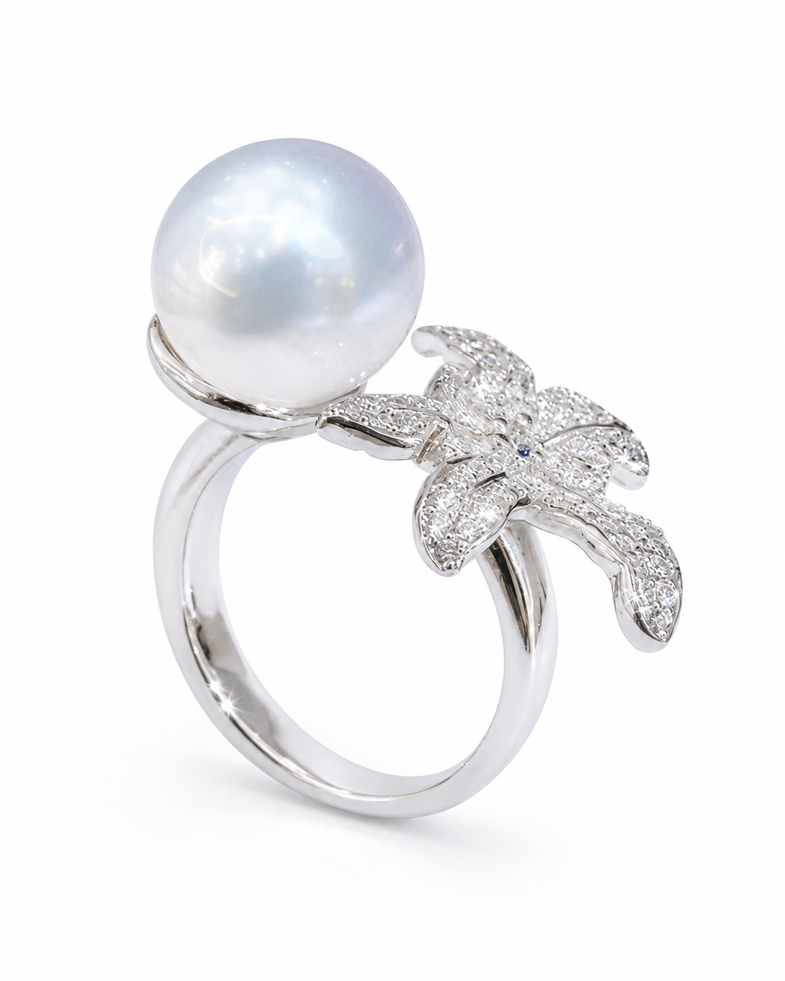 A silver ring featuring a large white pearl held in a setting with a pavé diamond flower and a tiny blue accent stone, shown on a clean white background.